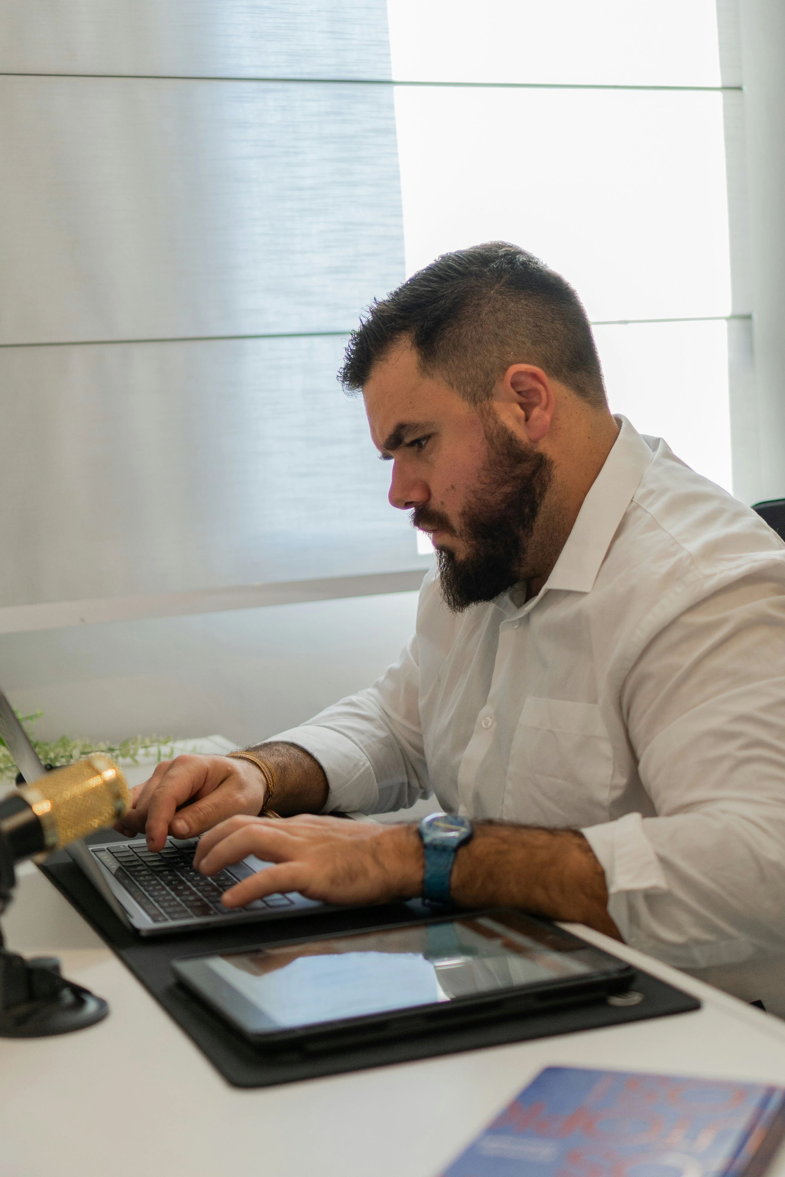 Man with beard and white shirt typing on a laptop at a desk with a tablet and book nearby.