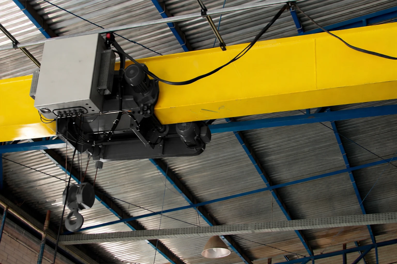 Yellow overhead crane with motor and hooks mounted inside an industrial building with blue steel roof trusses.