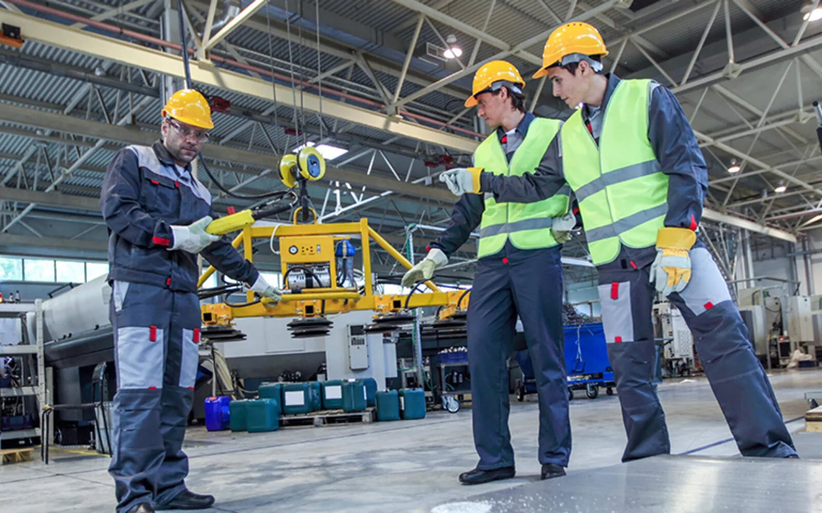 Three industrial workers wearing yellow helmets and safety gear operating a yellow lifting device inside a factory.