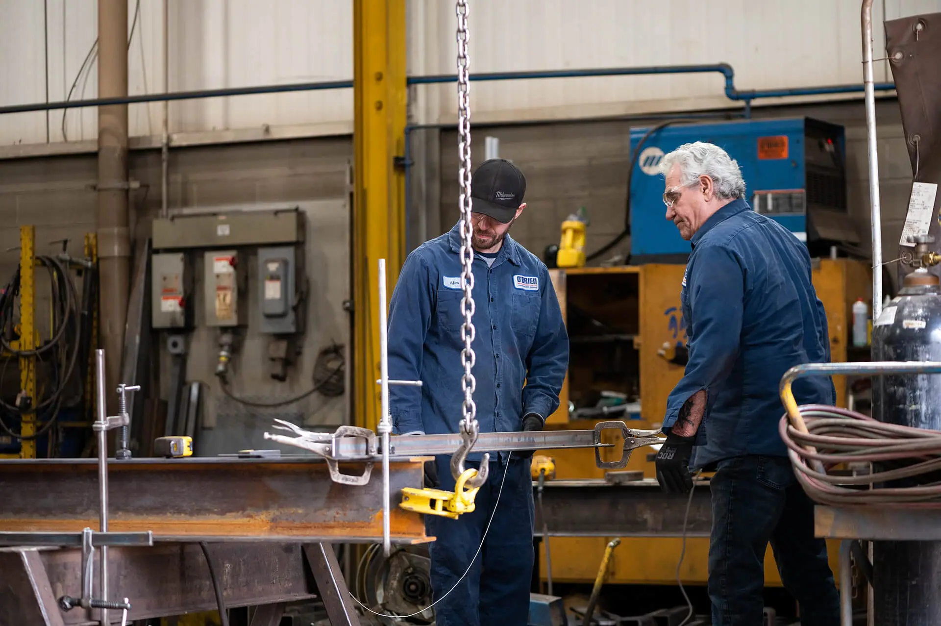 Two workers in blue uniforms operating heavy metal equipment in an industrial workshop.
