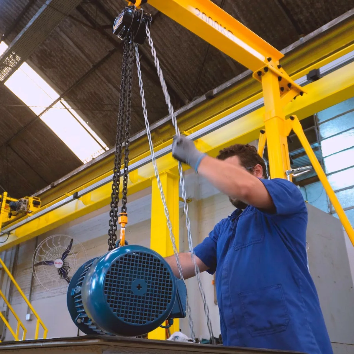 Worker in blue uniform operating a yellow industrial hoist lifting a large blue electric motor in a factory setting.