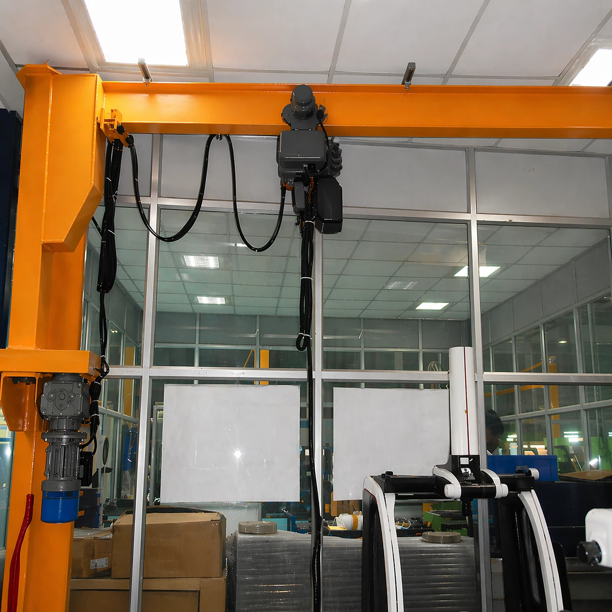 Indoor industrial setting with an orange overhead crane mechanism and packed boxes beneath glass windows.