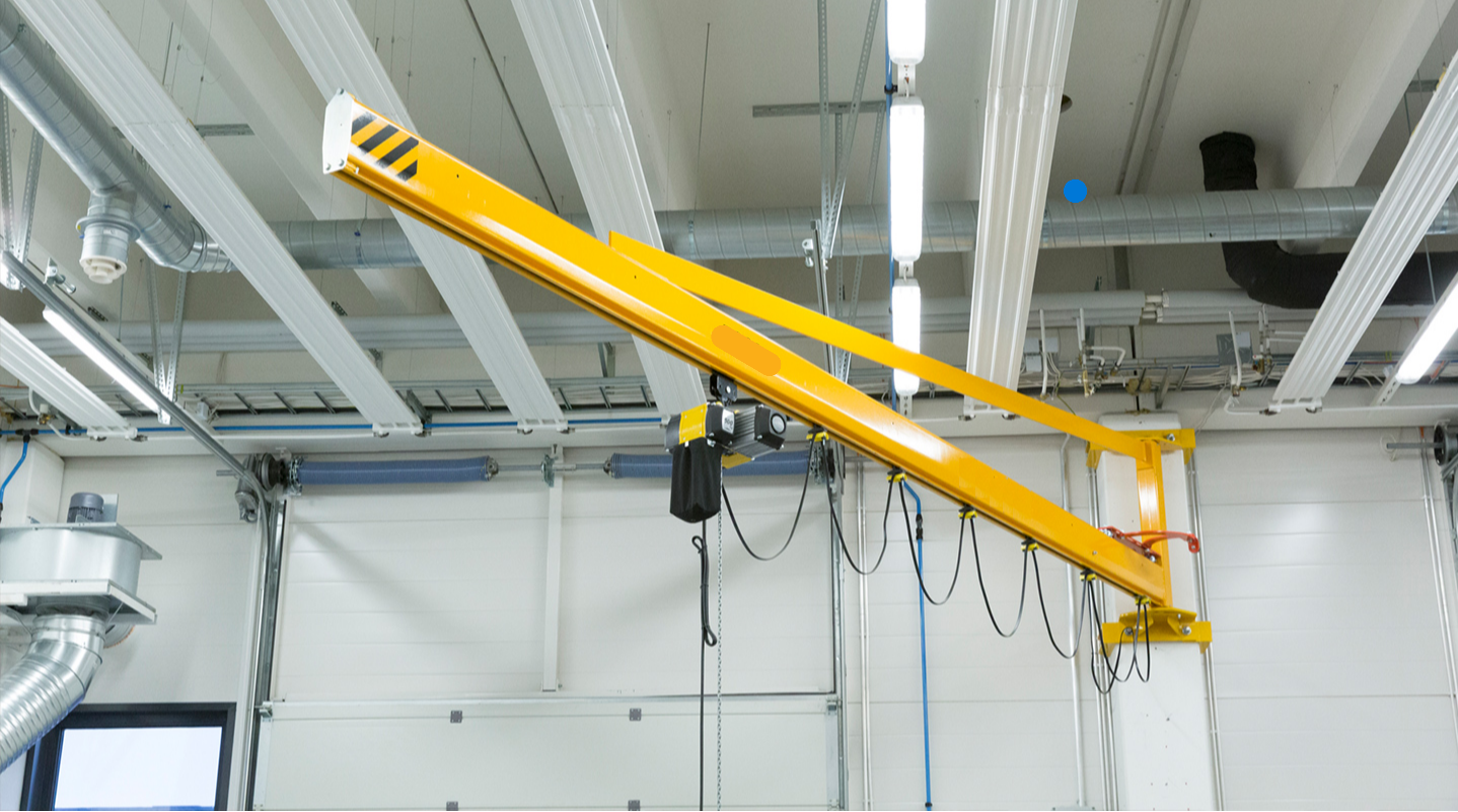 Yellow industrial jib crane mounted on a white wall inside a workshop with metal ductwork and fluorescent ceiling lights.