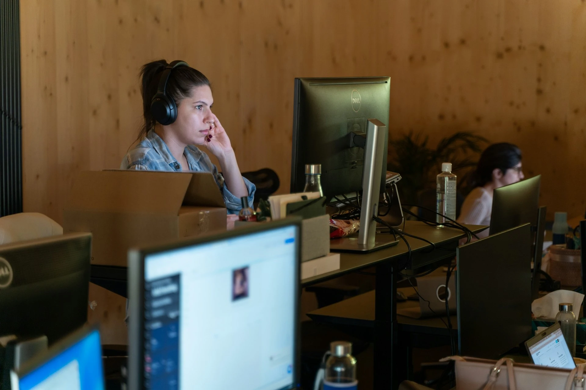 A woman wearing headphones works focused at a computer desk in an office environment with multiple monitors and another person working in the background.